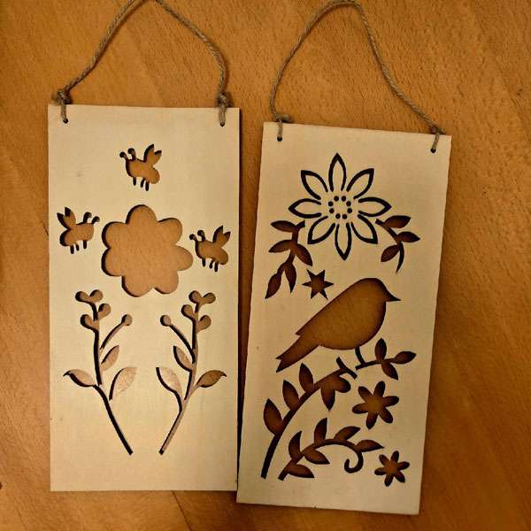 Set of two unfinished wooden hanging plaques with laser-cut floral, bee, and bird designs, displayed on a wooden table with jute hanging cords attached.